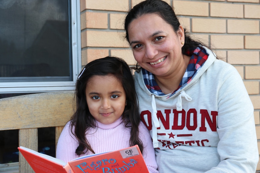 Indian mother smiling next to her young daughter, who is reading a book. 