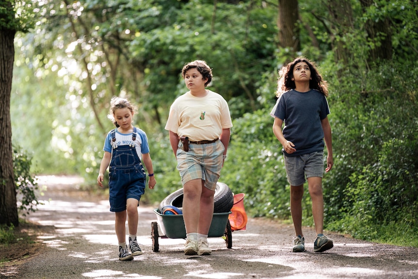 A small girl in overalls, a boy wheeling a tray and a boy with long hair walk on a dirt path surrounded by greenery.