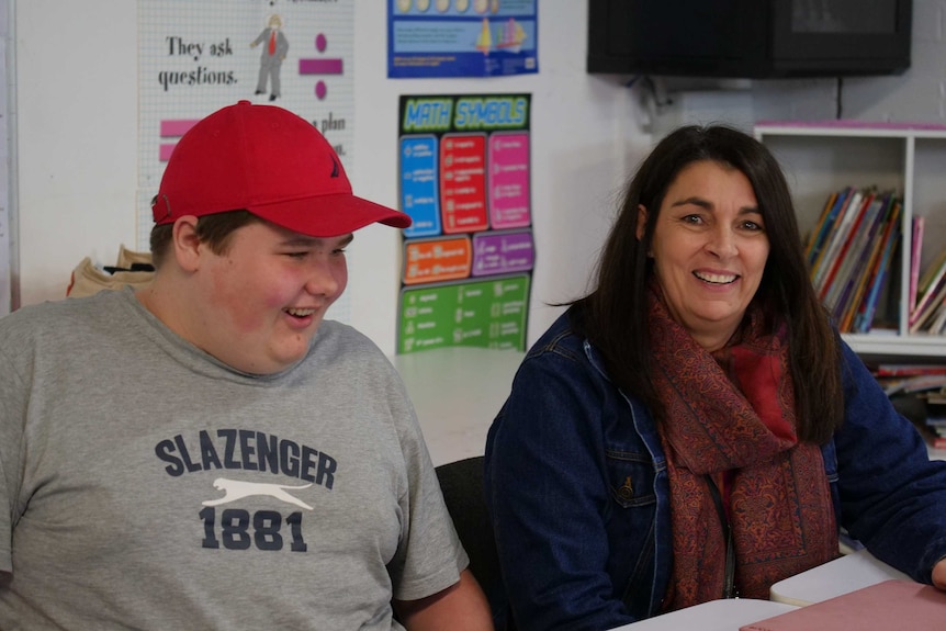 A teenage boy sits at a school desk laughing, next to a female teacher.