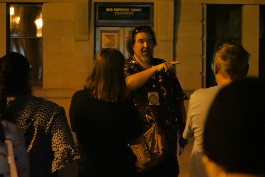 View of man, through people's shoulders, talking to people at night time pointing into distance