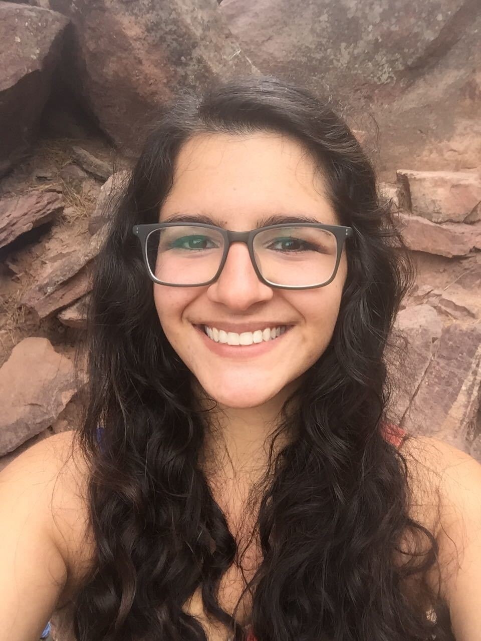 Un primer plano de una mujer joven con gafas y pelo largo y castaño que sonríe ampliamente, con un muro de rocas detrás de ella.