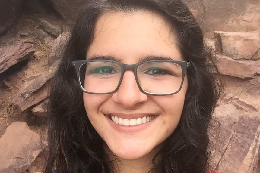 A close-up shot of young woman with glasses and long brown hair smiling broadly, with a wall of rocks behind her.