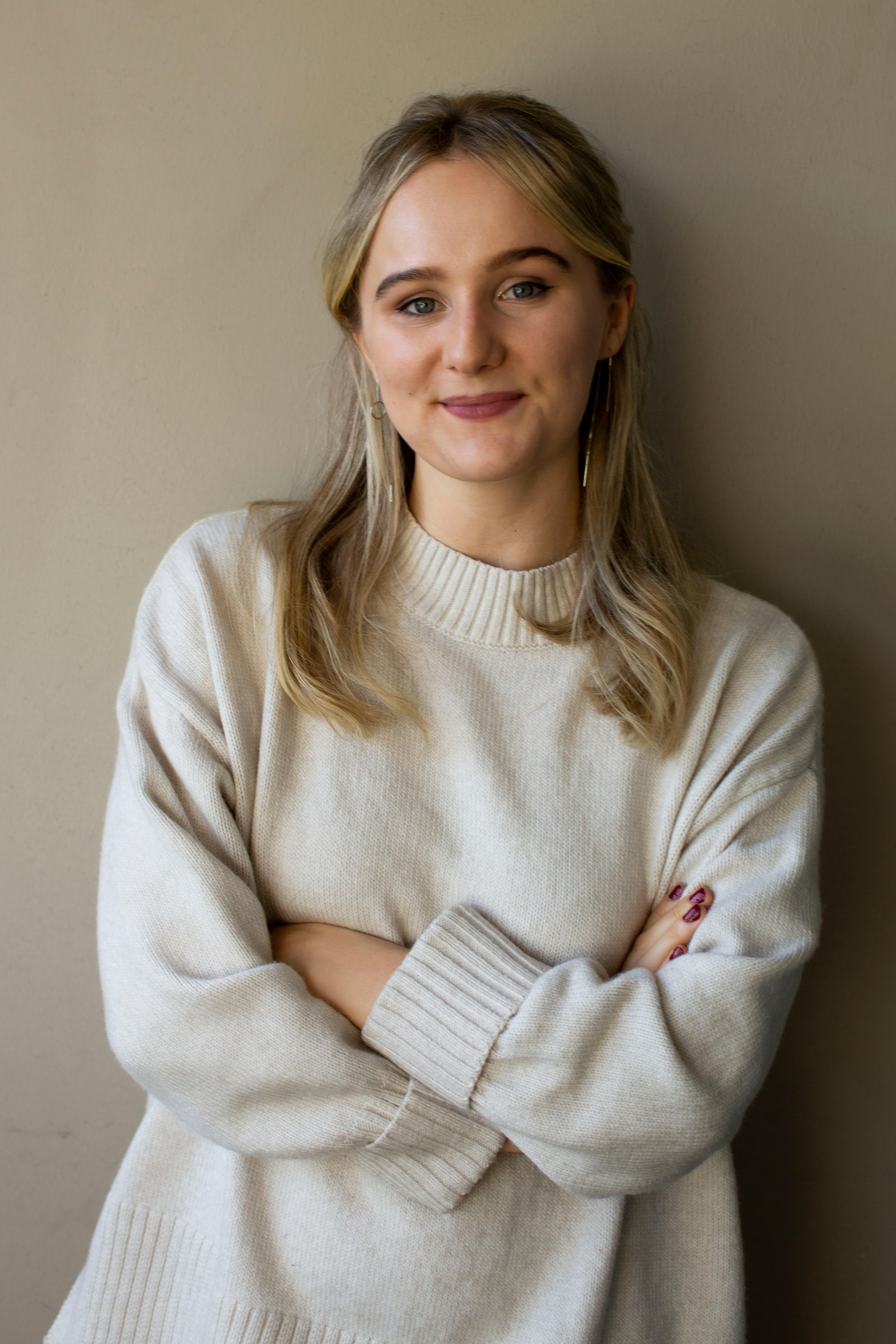A woman with blonde hair leans against a wall with her arms crossed, smiling.