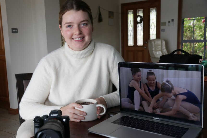 Woman having cup of tea, laptop open on right showing ballet dancers, camera in foreground on left 