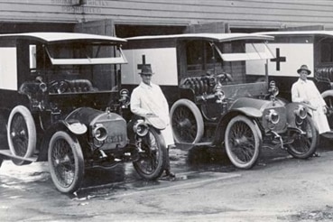 Black and white photo shows 1930s-era ambulances with crosses on the sides, with attendants beside them.
