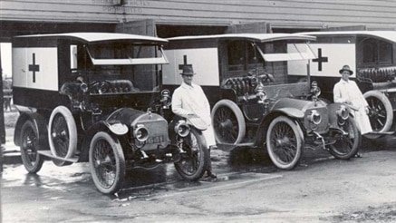 Black and white photo shows 1930s-era ambulances with crosses on the sides, with attendants beside them.