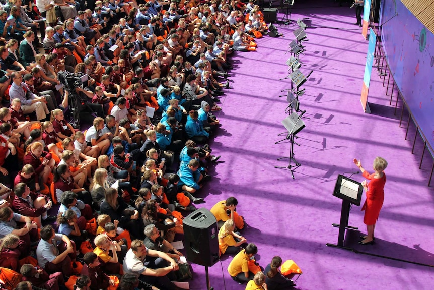 Hundreds of kids bathed in sunlight watch a lady in a red dress speak to them