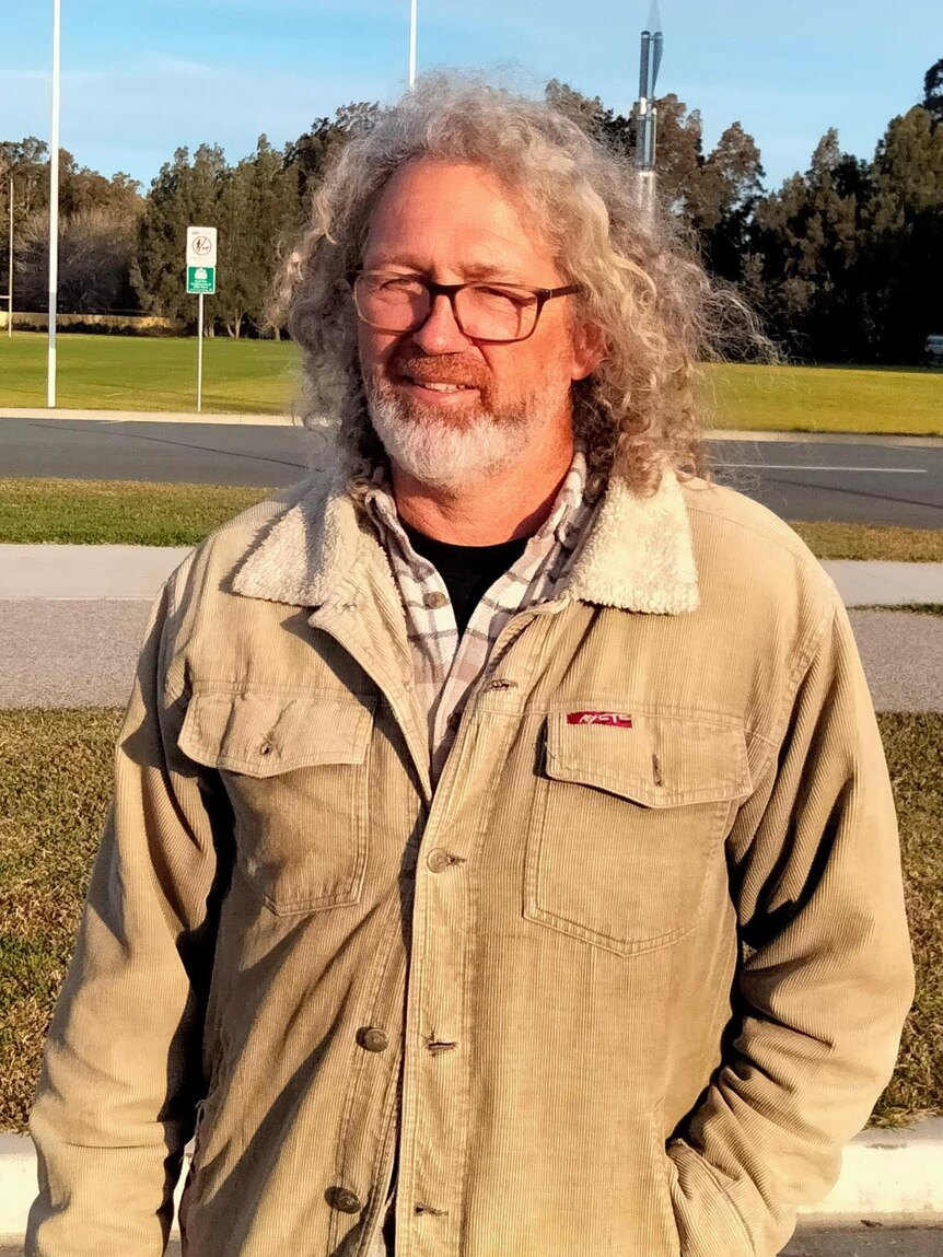 A man with curly grey hair and a beard, wearing glasses and a warm coat, smiling