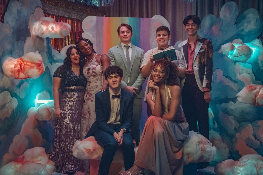 Seven young Heartstopper cast members in formal wear prom outfits pose for a photo in a prom-like setting