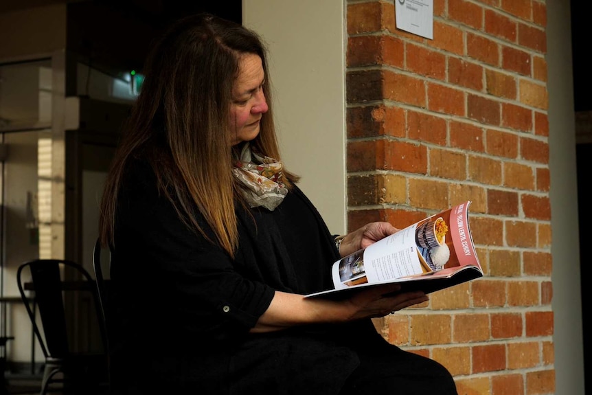 A woman sits looking at a recipe book in her hand.