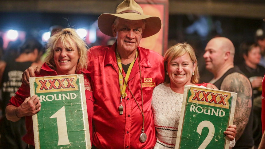 A man with an Akubra-style hat and a red shirt stands with his arms around two women holding signs saying Round 1 and Round 2.