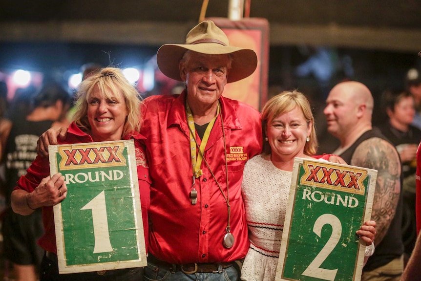 A man with an Akubra-style hat and a red shirt stands with his arms around two women holding signs saying Round 1 and Round 2.