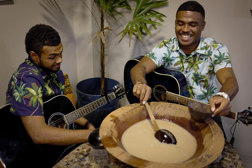 Two men in colourful shirts sit with guitars and a large bowl of kava. 
