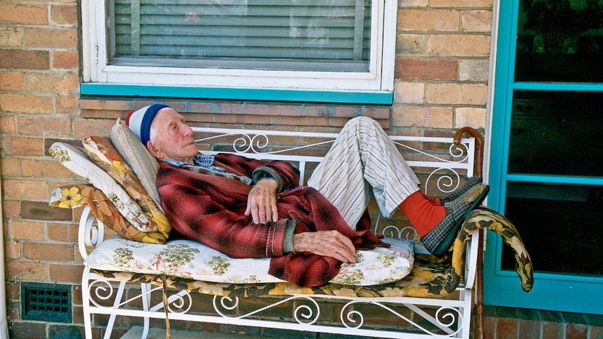 A man wearing a beanie and dressing gown lies on a bench on a suburban porch