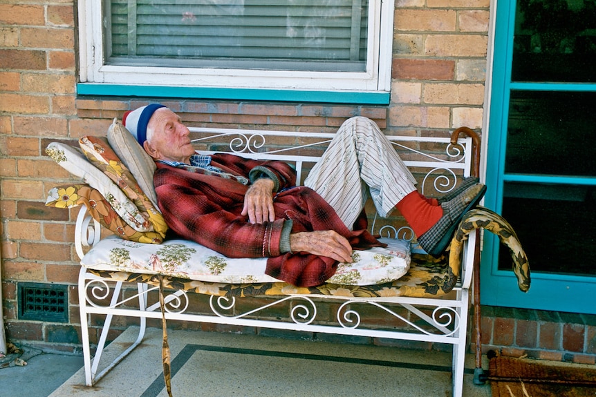 A man wearing a beanie and dressing gown lies on a bench on a suburban porch