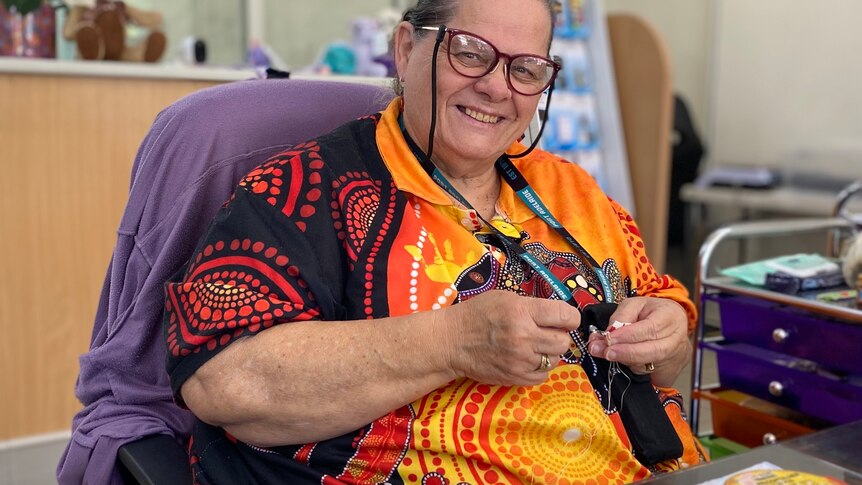 A woman smiles, she wears a black and orange top with indigenous art. She has short hair and sits in a chair, with crafts