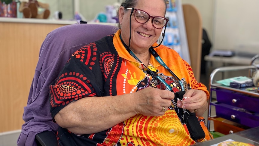 A woman smiles, she wears a black and orange top with indigenous art. She has short hair and sits in a chair, with crafts