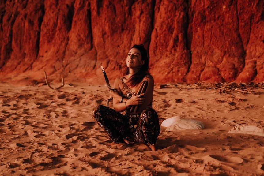 A young woman sitting cross-legged in sand with clapping sticks in her hand.