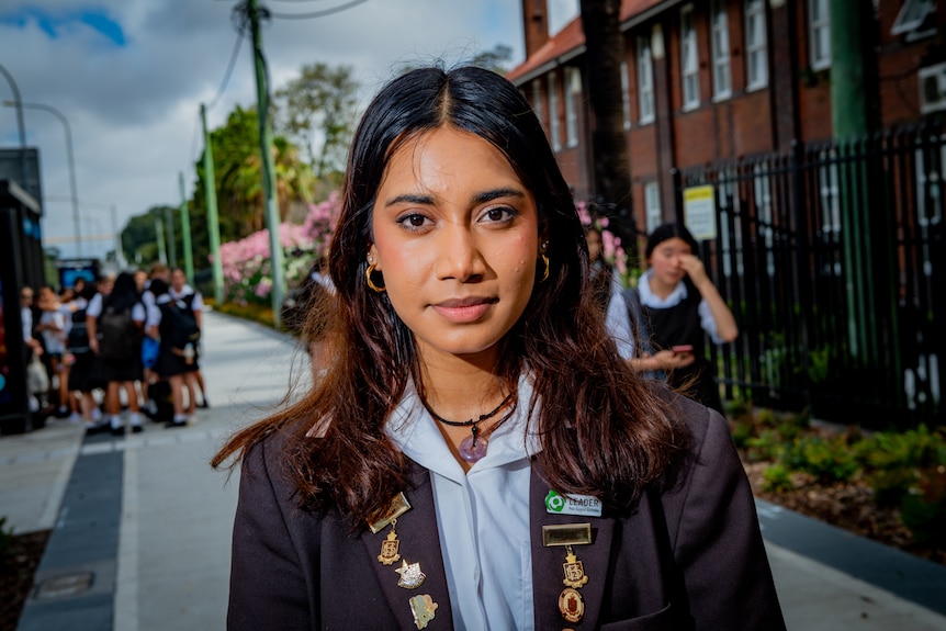 A girl with dark hair, wearing a blue school uniform stares into the camera, school girls blurred in background behind.