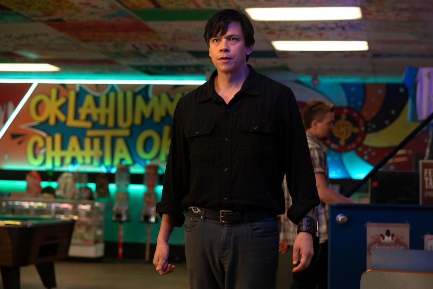 A TV still of Chaske Spencer, an Native American man in his late 40s, standing in a busy arcade.