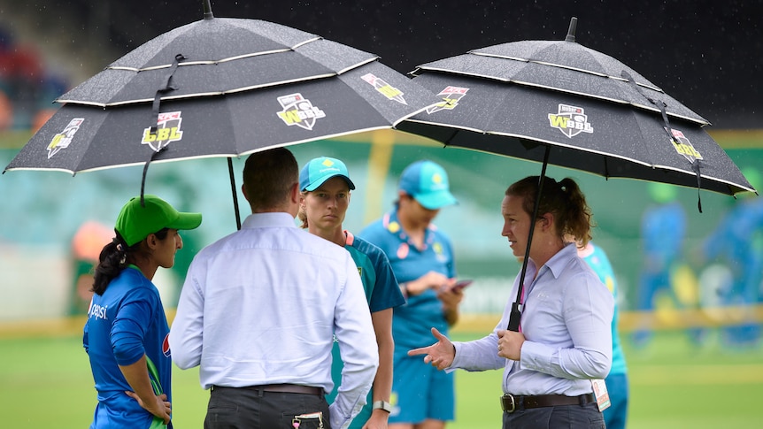 Two women's cricket captains stand on the ground with umpires holding umbrellas during a rain delay.