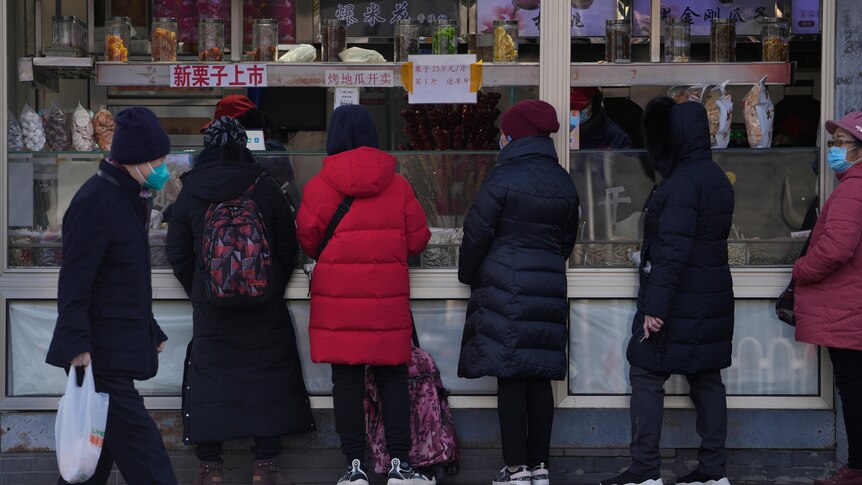 A resident wearing a face mask walks by women who line up to buy snack foods at a store in Beijing.