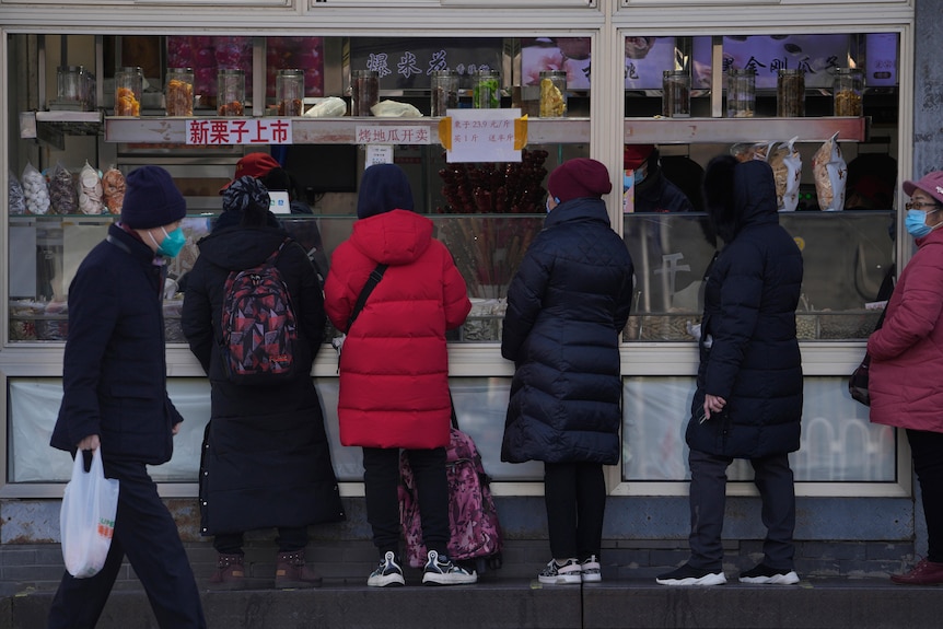 A resident wearing a face mask walks by women who line up to buy snack foods at a store in Beijing.