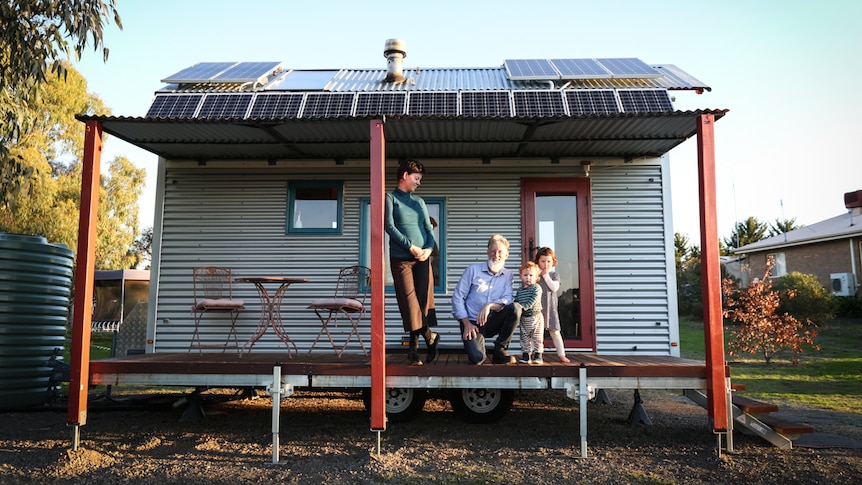 A family consisting of a man, woman and two young boys stands on the front verandah of their tiny house.