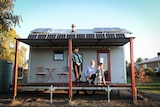 A family consisting of a man, woman and two young boys stands on the front verandah of their tiny house.