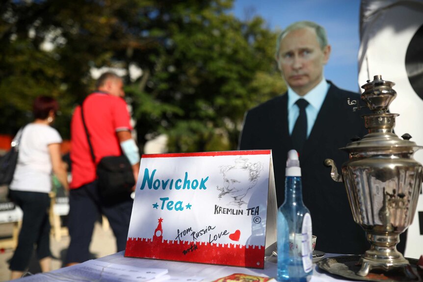 People pass a mock offer of "Novichok Tea" in front of a picture of Russian President Vladimir Putin
