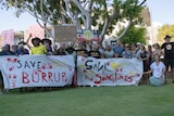 A large group of protesters stand in a park, holding up banners.