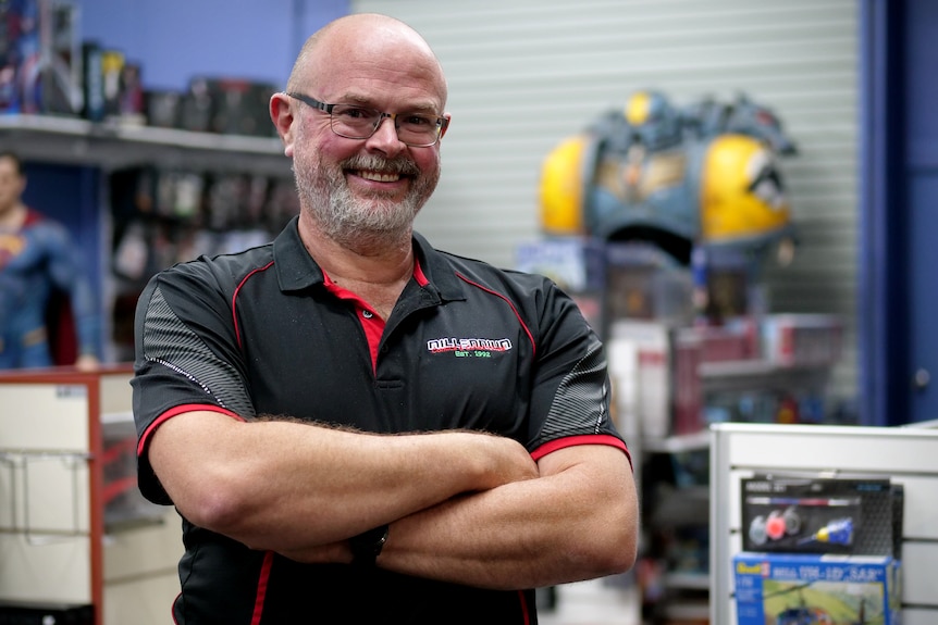 A bald man with facial hair and glasses smiles, he is wearing a black and red polo shirt
