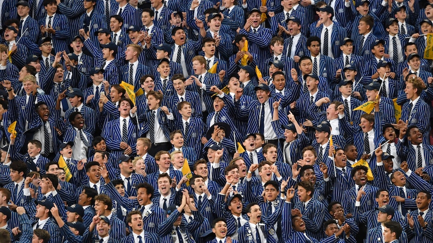 Dozens of high school boys students, in uniform, in a grandstand, cheering on their rugby team.