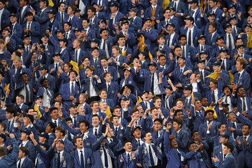 Dozens of high school boys students, in uniform, in a grandstand, cheering on their rugby team.