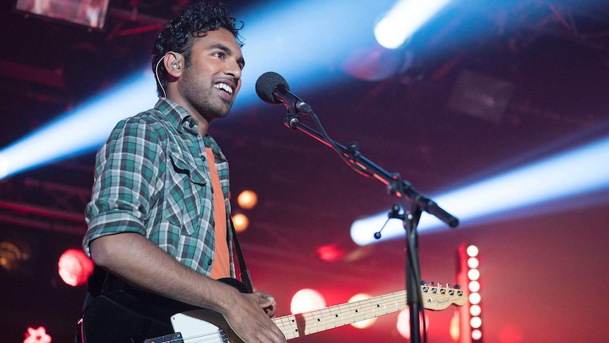Patel stands on a stage, guitar slung on shoulder, smiling out to the crowd.