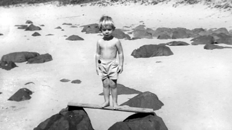 A black and white photo of artist Robert Brownhall as a boy aged 3, standing on a rock at the beach.