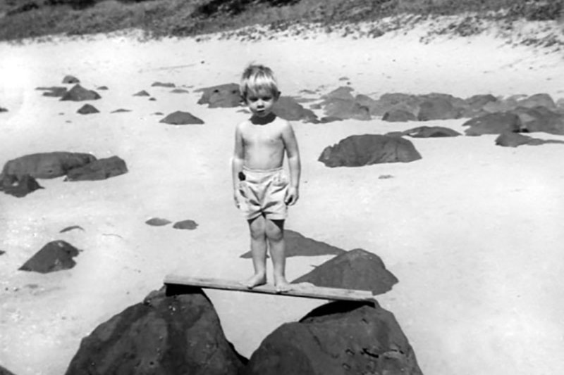 A black and white photo of artist Robert Brownhall as a boy aged 3, standing on a rock at the beach.