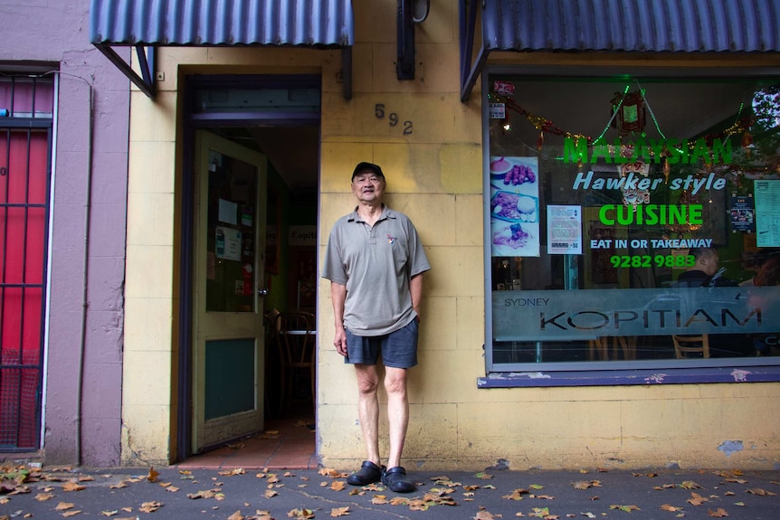 A man stands in front of a shop, wearing a grey shirt.