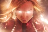 A close up portrait of Captain Marvel in costume with gold supernatural energy radiating from her eyes and around her head.
