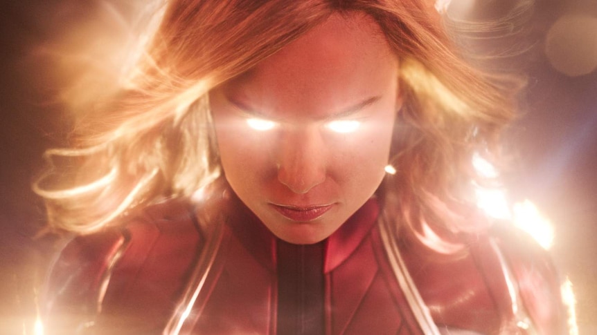 A close up portrait of Captain Marvel in costume with gold supernatural energy radiating from her eyes and around her head.