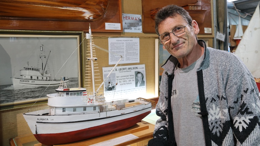 Man on right looking at camera standing next to wooden painted white ship model on shelf, picture of boat in background