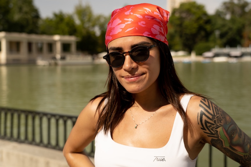 A young woman wearing a red and pink pattern bandana on her head and sunglasses stands beside a body of water