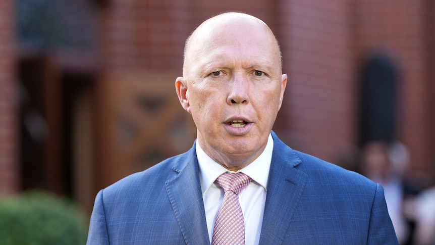 Peter Dutton wearing a blue suit and light pink tie standing in front of a brick building that is out of focus