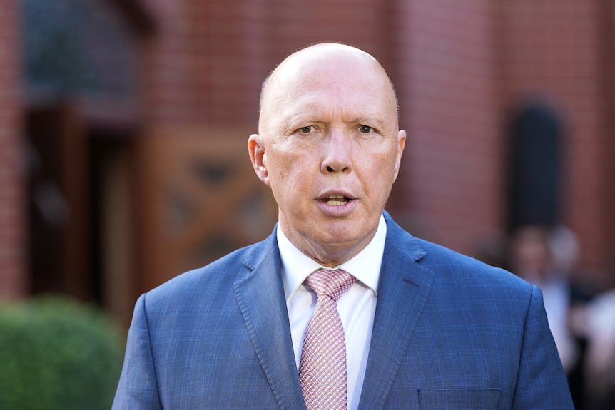 Peter Dutton wearing a blue suit and light pink tie standing in front of a brick building that is out of focus