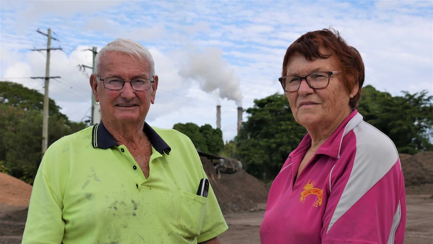 An older man and woman dressed in work outdoor work clothes stand outside. Smoke stacks and trees are visible in the background.