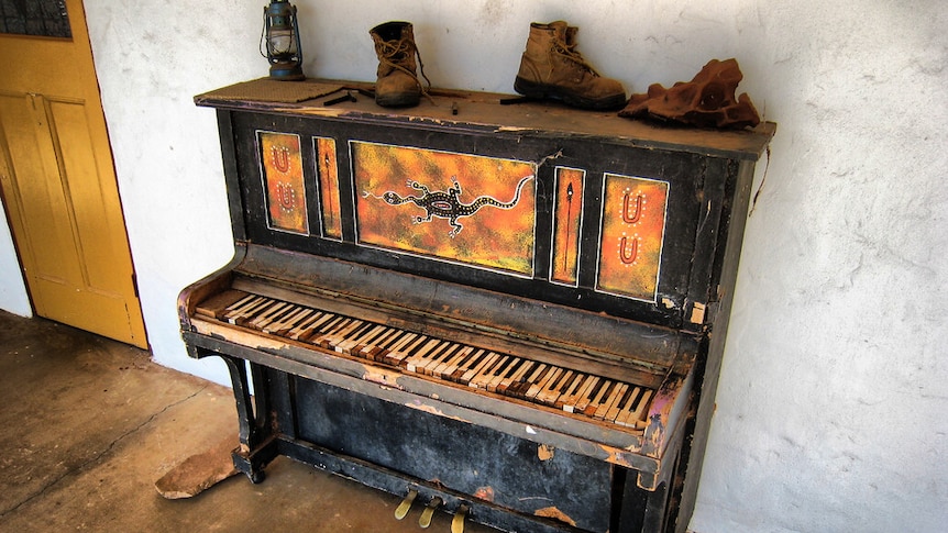 An old, broken upright piano, painted with Aboriginal artwork. Some old boots and a hurricane lantern sit on top.