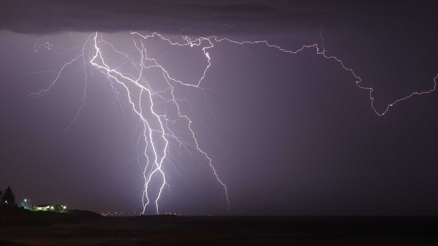 A huge fork of lightning cuts through the sky above a tiny lit-up building on the coast.