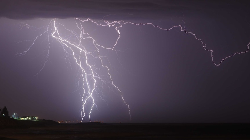 A huge fork of lightning cuts through the sky above a tiny lit-up building on the coast.