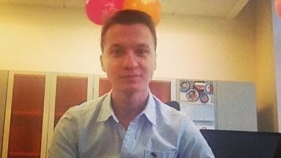 Aleksandr Ermakov wearing a light blue button up shirt, sitting at a desk with coloured balloons above him