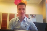 Aleksandr Ermakov wearing a light blue button up shirt, sitting at a desk with coloured balloons above him